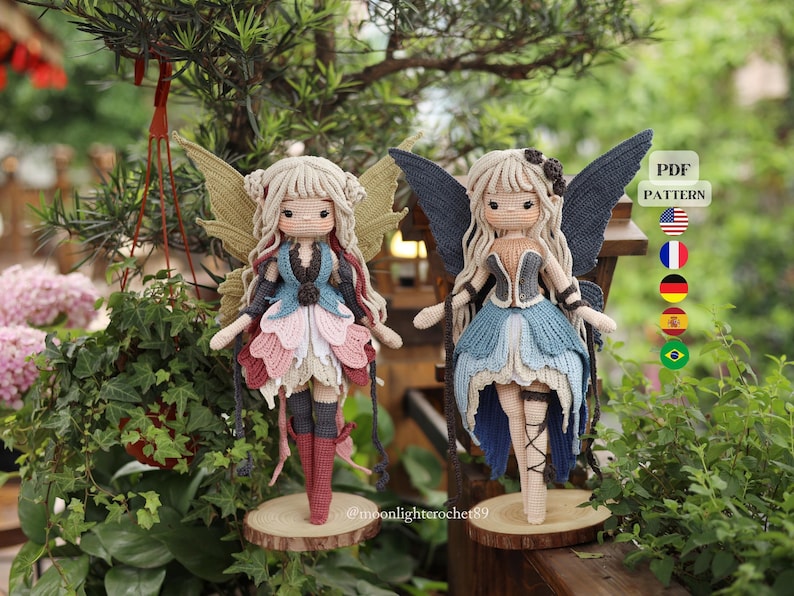 May include: Two handmade crochet fairy dolls. One doll has a blue, pink, and white dress, the other a blue and grey dress. Both dolls have detailed wings and hair, and stand on wooden bases. The background features lush green foliage.