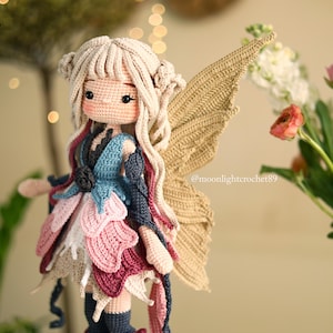 Peut inclure: Une poup&eacute;e f&eacute;e au crochet faite &agrave; la main avec des cheveux blonds et de grandes ailes beiges. La poup&eacute;e porte une robe superpos&eacute;e dans les tons de rose, de bleu et de bordeaux. La poup&eacute;e mesure environ 25 cm de haut. Le fond comprend des fleurs et un &eacute;clairage doux.
