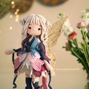 Peut inclure: Une poup&eacute;e f&eacute;e au crochet faite &agrave; la main, avec un teint beige clair, portant une robe multicolore avec des accents roses, bleus et blancs. La poup&eacute;e a des cheveux beiges, de grandes ailes et est pos&eacute;e devant un fond flou avec des fleurs.