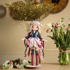 Peut inclure: Une poup&eacute;e de f&eacute;e au crochet faite &agrave; la main avec une peau claire, des cheveux blonds et des ailes dor&eacute;es. La poup&eacute;e porte une robe bleue, rose et blanche avec des bottes assorties. La f&eacute;e est debout sur une base en bois, avec des fleurs en arri&egrave;re-plan.