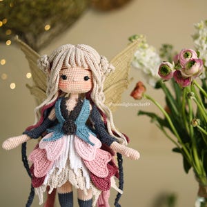 Peut inclure: Poup&eacute;e f&eacute;e au crochet faite &agrave; la main, avec des cheveux bruns clairs, des ailes et une robe multicouche dans les tons de bleu, rose et blanc. La poup&eacute;e a un visage d&eacute;taill&eacute; et se trouve devant un vase de fleurs.