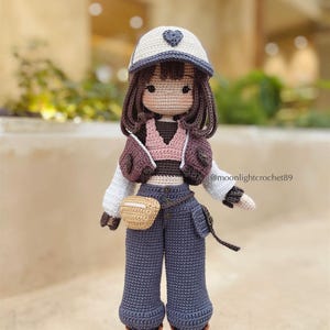 Puede incluir: Una mu&ntilde;eca de crochet que lleva una gorra de b&eacute;isbol blanca y gris con un coraz&oacute;n, una chaqueta marr&oacute;n, una camiseta rosa, pantalones vaqueros azules, un cintur&oacute;n marr&oacute;n, una bolsa marr&oacute;n y botas marrones. La mu&ntilde;eca tiene el pelo marr&oacute;n y una cara de crochet.