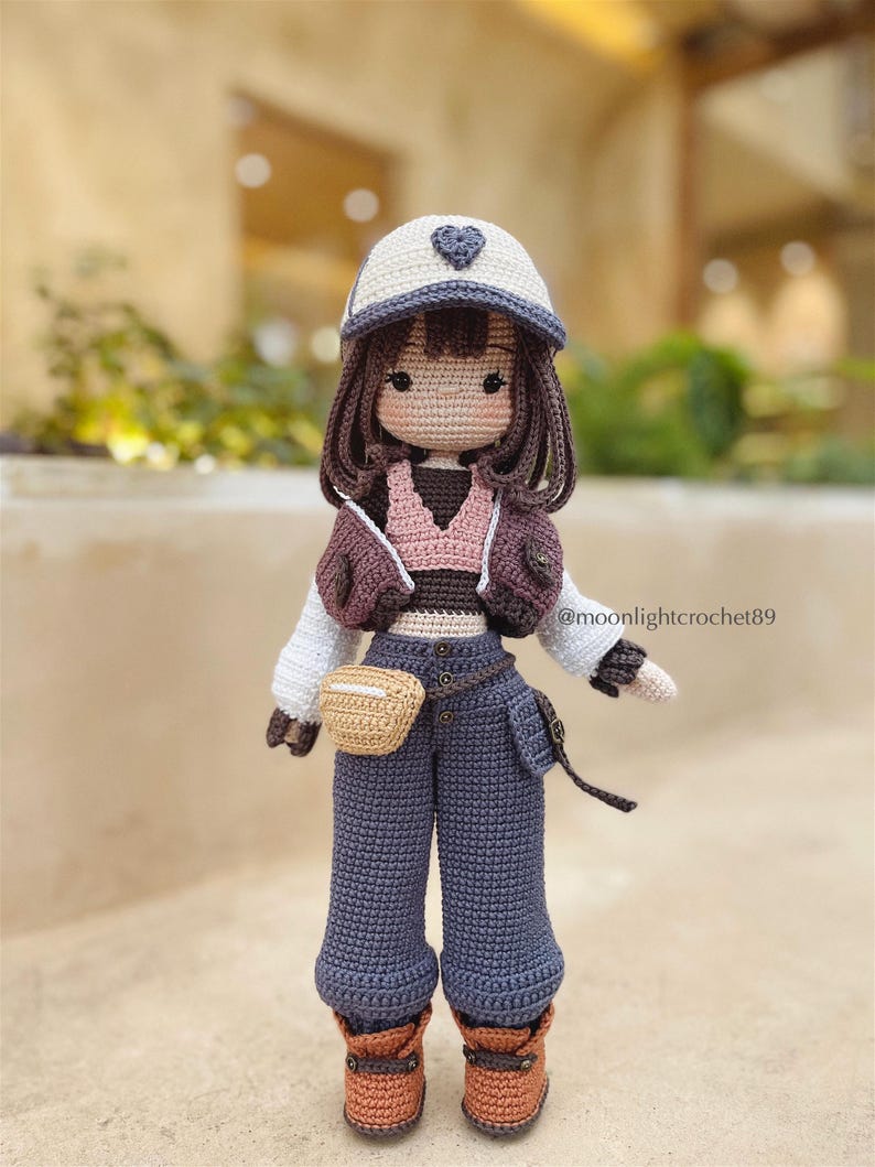 Puede incluir: Una mu&ntilde;eca de crochet que lleva una gorra de b&eacute;isbol blanca y gris con un coraz&oacute;n, una chaqueta marr&oacute;n, una camiseta rosa, pantalones vaqueros azules, un cintur&oacute;n marr&oacute;n, una bolsa marr&oacute;n y botas marrones. La mu&ntilde;eca tiene el pelo marr&oacute;n y una cara de crochet.