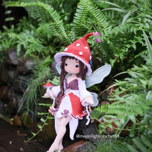 May include: A handmade crochet fairy doll with a red and white mushroom cap hat and wings. The doll is wearing a white dress with red accents and is surrounded by greenery. The doll is sitting on a wooden surface. The text "@moonlightcrochet89" is visible.