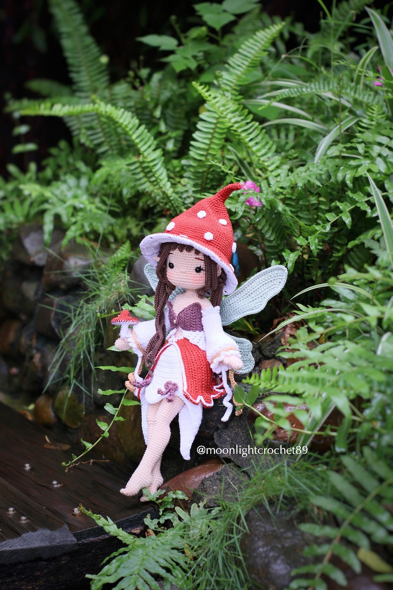 May include: A handmade crochet fairy doll with a red and white mushroom cap hat and wings. The doll is wearing a white dress with red accents and is surrounded by greenery. The doll is sitting on a wooden surface. The text "@moonlightcrochet89" is visible.