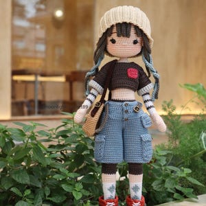May include: A crocheted doll wearing a brown shirt with a red heart, blue jeans, a beige hat, and brown boots. The doll is holding a brown bag.
