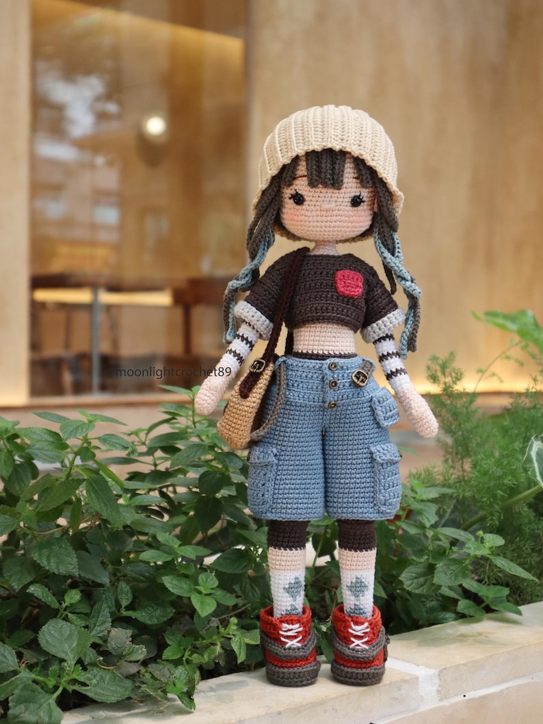 May include: A crocheted doll wearing a brown shirt with a red heart, blue jeans, a beige hat, and brown boots. The doll is holding a brown bag.
