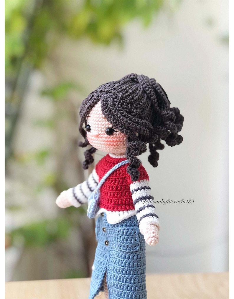 May include: A crocheted doll with brown hair, a red and white striped shirt, a red sweater, a blue denim skirt, and a small brown bag. The doll is standing on a wooden surface. @moonlightcrochet89