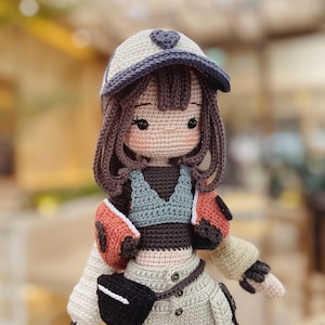 Puede incluir: Una mu&ntilde;eca de crochet que lleva una gorra de b&eacute;isbol blanca y morada, una chaqueta marr&oacute;n y naranja, un top gris y pantalones beige con un cintur&oacute;n negro y una ri&ntilde;onera negra. La mu&ntilde;eca tiene el pelo marr&oacute;n y la cara rosa con ojos marrones.