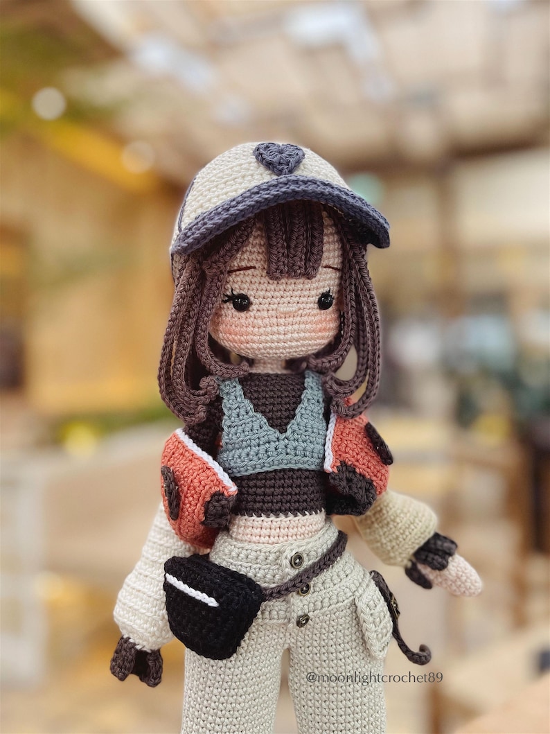 Puede incluir: Una mu&ntilde;eca de crochet que lleva una gorra de b&eacute;isbol blanca y morada, una chaqueta marr&oacute;n y naranja, un top gris y pantalones beige con un cintur&oacute;n negro y una ri&ntilde;onera negra. La mu&ntilde;eca tiene el pelo marr&oacute;n y la cara rosa con ojos marrones.