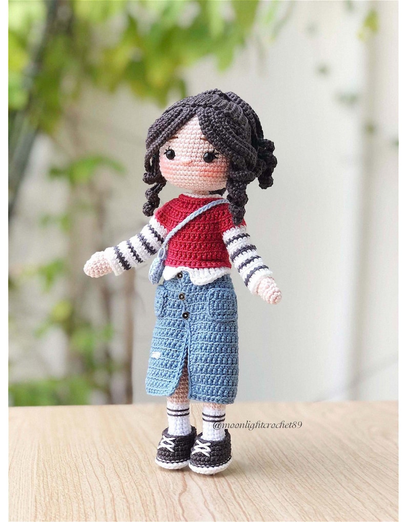 May include: Crocheted doll with black hair, wearing a red and white striped shirt, a blue denim skirt, and white socks with black shoes. The doll is holding a small brown bag.