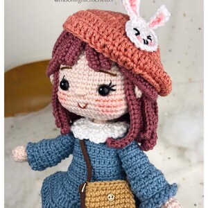 May include: Crocheted doll with pink hair, wearing a blue dress, a brown purse, and a pink beret with a white bunny applique. The doll has a white collar and brown eyes. The doll is holding a brown purse with a gold button.