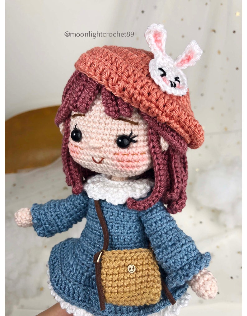 May include: Crocheted doll with pink hair, wearing a blue dress, a brown purse, and a pink beret with a white bunny applique. The doll has a white collar and brown eyes. The doll is holding a brown purse with a gold button.