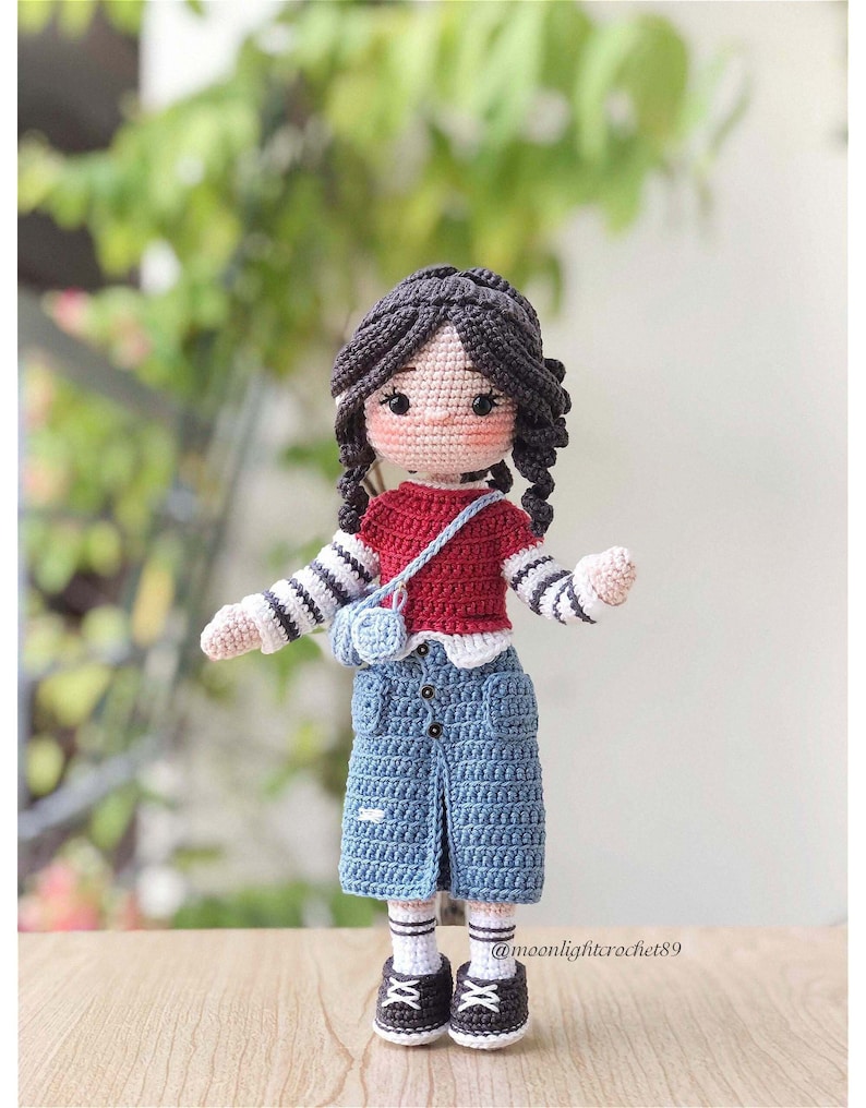 May include: A crocheted doll with brown hair, wearing a red and white striped shirt, a blue denim skirt, and white socks with black shoes. The doll is holding a small red bag with a white heart on it.