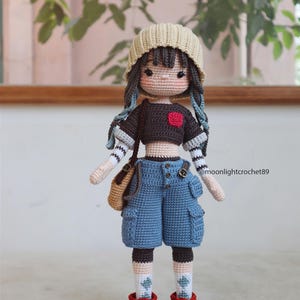 May include: Crocheted doll with brown hair, wearing a beige beanie, a black and white striped shirt, blue denim shorts, and red and black sneakers. The doll is holding a brown bag.