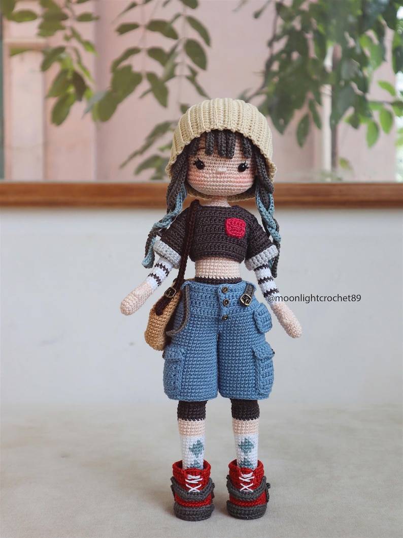 May include: Crocheted doll with brown hair, wearing a beige beanie, a black and white striped shirt, blue denim shorts, and red and black sneakers. The doll is holding a brown bag.