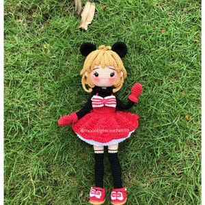 K&ouml;nnte beinhalten: Eine geh&auml;kelte Puppe mit blonden Haaren, die ein rotes Kleid mit wei&szlig;en Verzierungen, schwarze Str&uuml;mpfe und rote Schuhe mit wei&szlig;en Schleifen tr&auml;gt. Die Puppe hat schwarze M&auml;useohren und h&auml;lt in einer Hand eine rote M&uuml;tze. Die Puppe sitzt auf einem Bett aus gr&uuml;nem Gras. @moonlightcrochet89