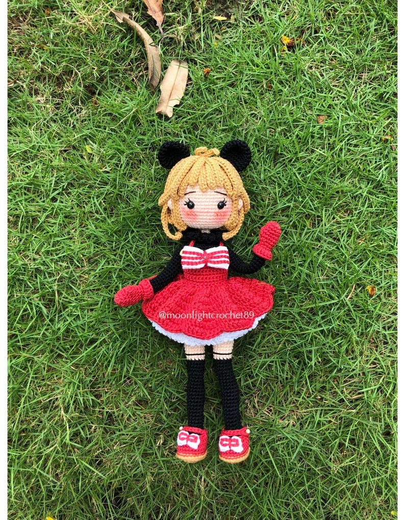 K&ouml;nnte beinhalten: Eine geh&auml;kelte Puppe mit blonden Haaren, die ein rotes Kleid mit wei&szlig;en Verzierungen, schwarze Str&uuml;mpfe und rote Schuhe mit wei&szlig;en Schleifen tr&auml;gt. Die Puppe hat schwarze M&auml;useohren und h&auml;lt in einer Hand eine rote M&uuml;tze. Die Puppe sitzt auf einem Bett aus gr&uuml;nem Gras. @moonlightcrochet89
