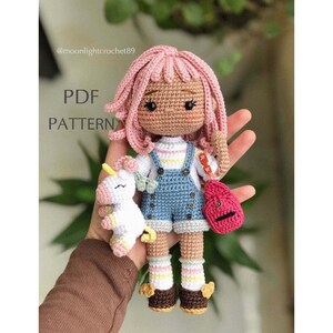 Puede incluir: Mu&ntilde;eca de crochet con pelo rosa, vistiendo un vestido de mezclilla azul, una camisa a rayas y sosteniendo un unicornio blanco. La mu&ntilde;eca lleva zapatos marrones con detalles amarillos y tiene una mochila roja.