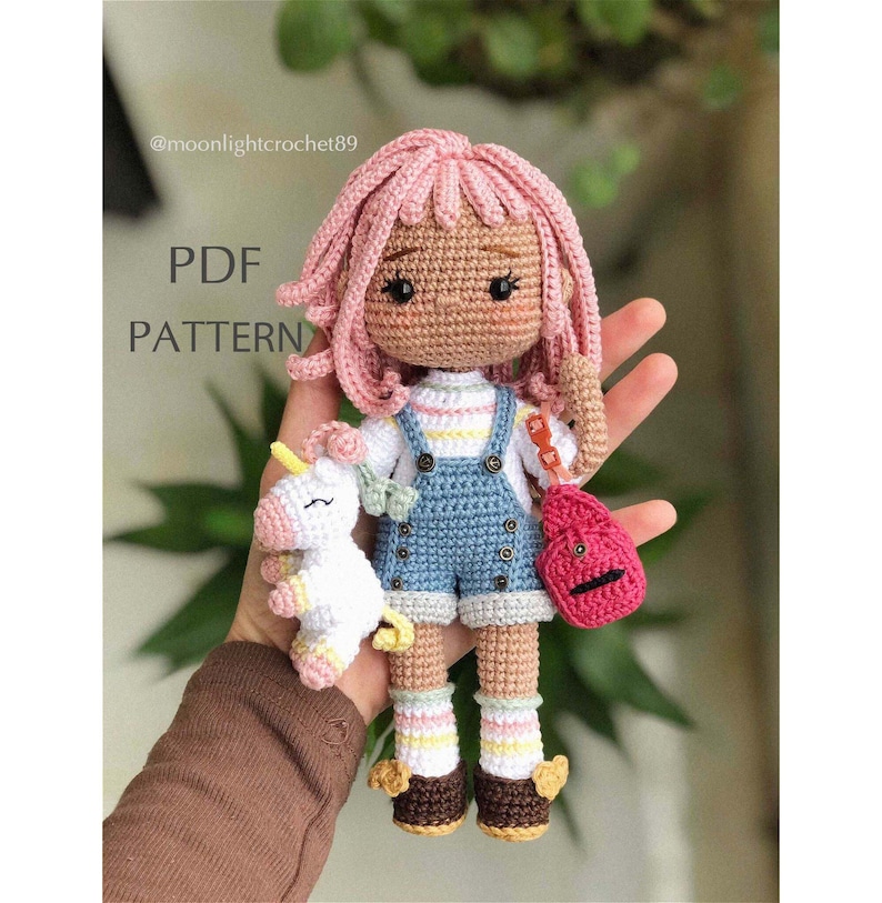 Puede incluir: Mu&ntilde;eca de crochet con pelo rosa, vistiendo un vestido de mezclilla azul, una camisa a rayas y sosteniendo un unicornio blanco. La mu&ntilde;eca lleva zapatos marrones con detalles amarillos y tiene una mochila roja.