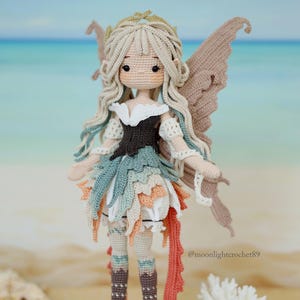 Puede incluir: Una mu&ntilde;eca de ganchillo hecha a mano con largo cabello rubio, un corpi&ntilde;o oscuro y una falda de capas en tonos verde azulado, melocot&oacute;n y blanco. La mu&ntilde;eca tiene grandes ojos marrones, alas intrincadas y botas altas marrones. El texto @moonlightcrochet89 es visible.