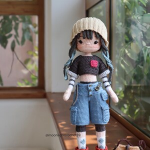 May include: A crocheted doll wearing a beige beanie, a black and brown striped shirt, blue denim shorts, red and brown shoes, and white socks with green accents. The doll is holding a brown crocheted bag with a brown strap.
