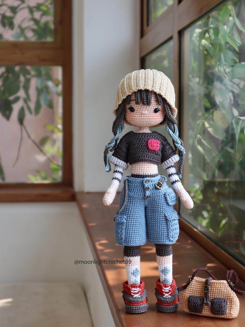 May include: A crocheted doll wearing a beige beanie, a black and brown striped shirt, blue denim shorts, red and brown shoes, and white socks with green accents. The doll is holding a brown crocheted bag with a brown strap.