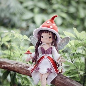 May include: A handmade crochet fairy doll with a red and white mushroom cap hat, white dress with red trim, and gray wings. The doll is sitting on a wooden branch with a green leafy background. The text "@moonlightcrochet89" is visible.
