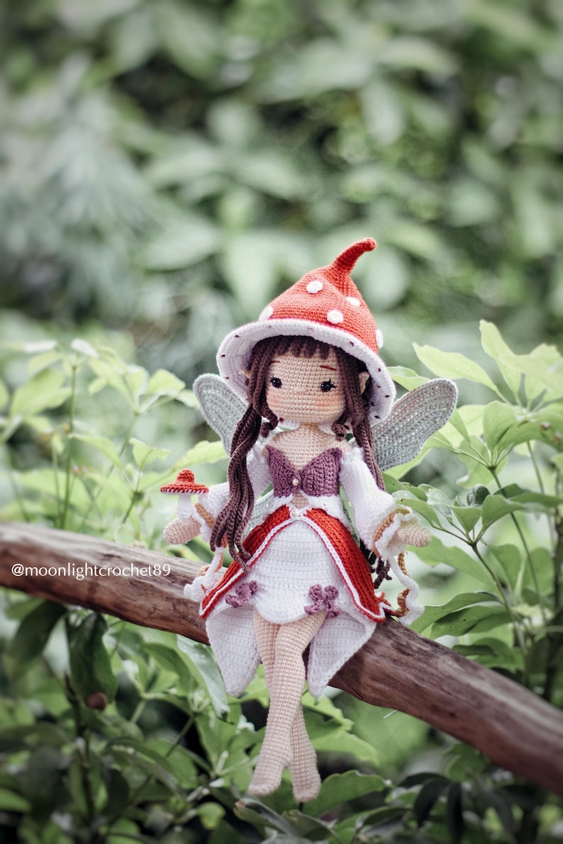 May include: A handmade crochet fairy doll with a red and white mushroom cap hat, white dress with red trim, and gray wings. The doll is sitting on a wooden branch with a green leafy background. The text "@moonlightcrochet89" is visible.