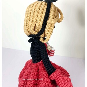K&ouml;nnte beinhalten: H&auml;kelpuppe mit schwarzen und wei&szlig;en Ohren, einem schwarzen Oberteil, einem roten Rock und einem wei&szlig;en Saum. Die Puppe h&auml;lt eine rot-wei&szlig; gestreifte H&auml;keltasche.