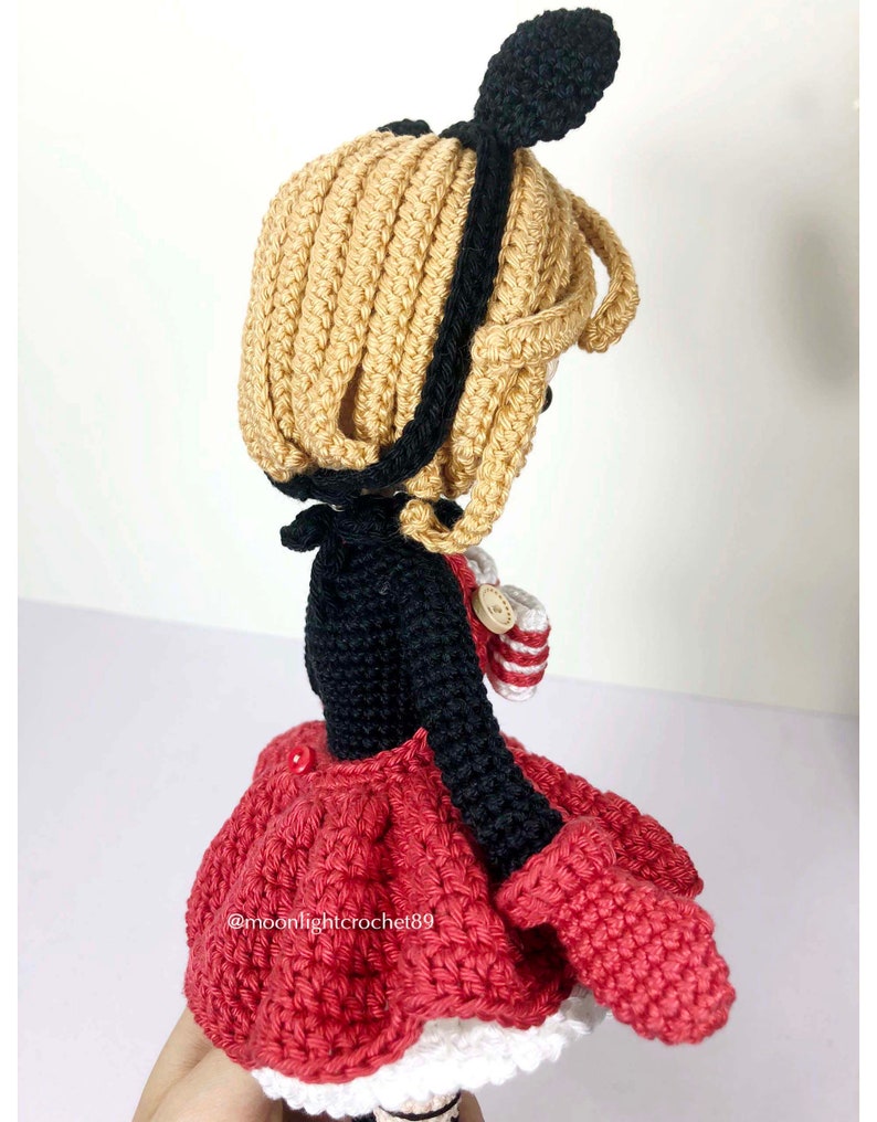 K&ouml;nnte beinhalten: H&auml;kelpuppe mit schwarzen und wei&szlig;en Ohren, einem schwarzen Oberteil, einem roten Rock und einem wei&szlig;en Saum. Die Puppe h&auml;lt eine rot-wei&szlig; gestreifte H&auml;keltasche.