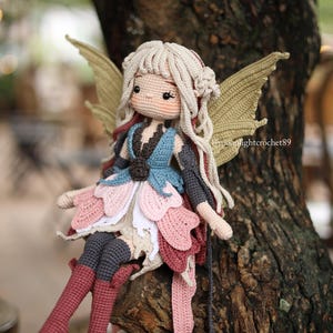 May include: A handmade crochet fairy doll with blonde hair, rosy cheeks, and large, detailed wings. The doll is wearing a layered dress in shades of pink, blue, and white, and is seated on a tree branch. The doll's boots are a deep red.