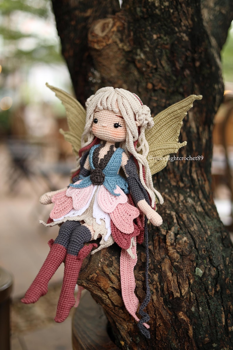 May include: A handmade crochet fairy doll with blonde hair, rosy cheeks, and large, detailed wings. The doll is wearing a layered dress in shades of pink, blue, and white, and is seated on a tree branch. The doll's boots are a deep red.