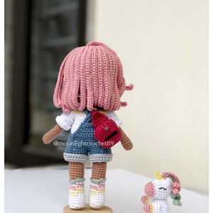 Puede incluir: Una mu&ntilde;eca de crochet con pelo rosa, que lleva una camisa blanca, un mono azul y una mochila roja. La mu&ntilde;eca est&aacute; de pie sobre una base de madera. Un peque&ntilde;o unicornio de crochet est&aacute; sentado en la mesa junto a la mu&ntilde;eca. El nombre de usuario de Instagram de la mu&ntilde;eca es @moonlightcrochet89.