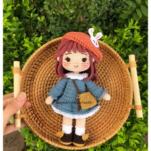 May include: A crocheted doll wearing a blue dress, a brown hat with a white bunny ear, and brown shoes. The doll is holding a brown purse and is sitting in a wicker basket.