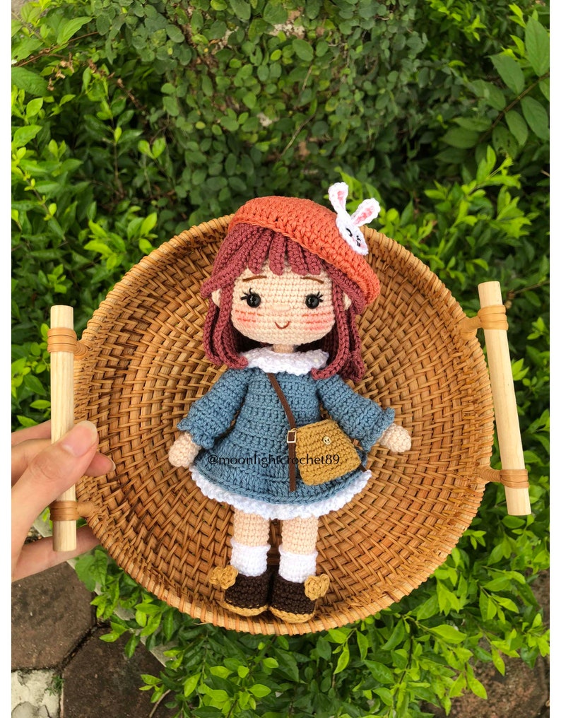 May include: A crocheted doll wearing a blue dress, a brown hat with a white bunny ear, and brown shoes. The doll is holding a brown purse and is sitting in a wicker basket.