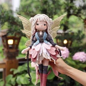 May include: A handmade crochet fairy doll with a light skin tone, blonde hair, and large, tan wings. The doll wears a blue, pink, and white dress with matching boots. The doll is held in front of a blurred green background.