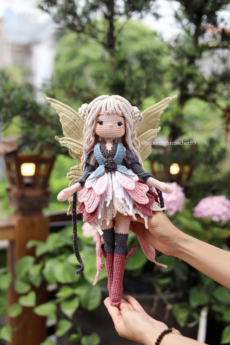 May include: A handmade crochet fairy doll with a light skin tone, blonde hair, and large, tan wings. The doll wears a blue, pink, and white dress with matching boots. The doll is held in front of a blurred green background.