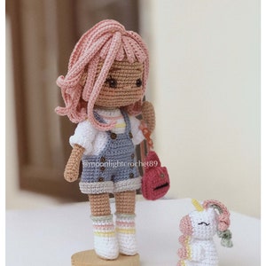 Puede incluir: Una mu&ntilde;eca de crochet con cabello rosado, un atuendo azul y blanco y una bolsa roja se encuentra sobre una base de madera. Un peque&ntilde;o unicornio de crochet blanco con detalles rosas y amarillos se sienta en el suelo junto a la mu&ntilde;eca. El nombre de usuario de Instagram de la mu&ntilde;eca es @moonlightcrochet89.