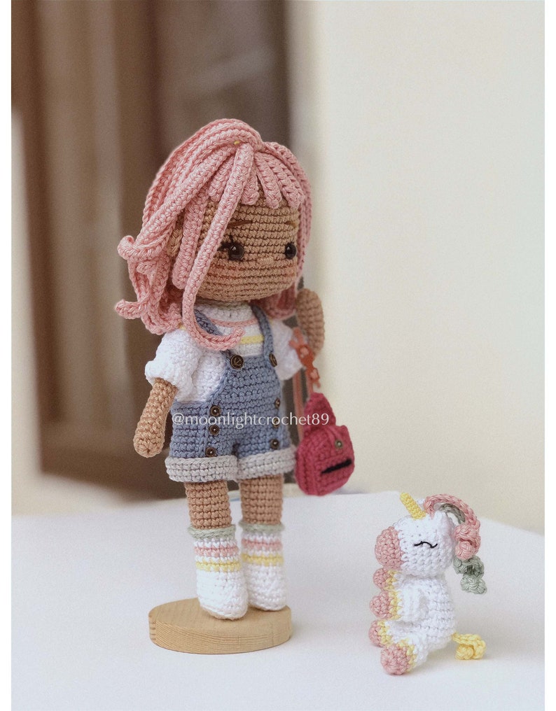 Puede incluir: Una mu&ntilde;eca de crochet con cabello rosado, un atuendo azul y blanco y una bolsa roja se encuentra sobre una base de madera. Un peque&ntilde;o unicornio de crochet blanco con detalles rosas y amarillos se sienta en el suelo junto a la mu&ntilde;eca. El nombre de usuario de Instagram de la mu&ntilde;eca es @moonlightcrochet89.