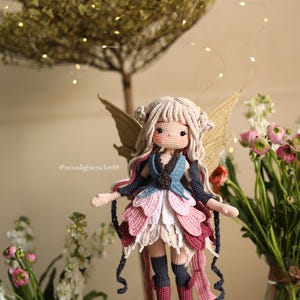 Peut inclure: Une poup&eacute;e f&eacute;e au crochet faite &agrave; la main, avec des cheveux blonds, des ailes dor&eacute;es et une robe color&eacute;e dans les tons de rose, bleu et blanc. La poup&eacute;e est devant un arri&egrave;re-plan flou avec des fleurs et un arbre. Le compte Instagram du cr&eacute;ateur de la poup&eacute;e, @moonlightcrochet89, est visible.
