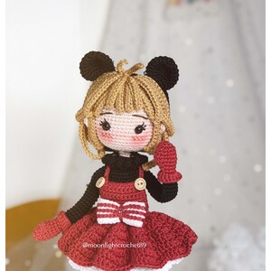 K&ouml;nnte beinhalten: Eine geh&auml;kelte Puppe mit blonden Haaren, die ein rotes Kleid mit einer wei&szlig;en Schleife, schwarze Schuhe und schwarze und rote F&auml;ustlinge tr&auml;gt. Die Puppe hat schwarze M&auml;useohren und einen schwarzen Pullover.