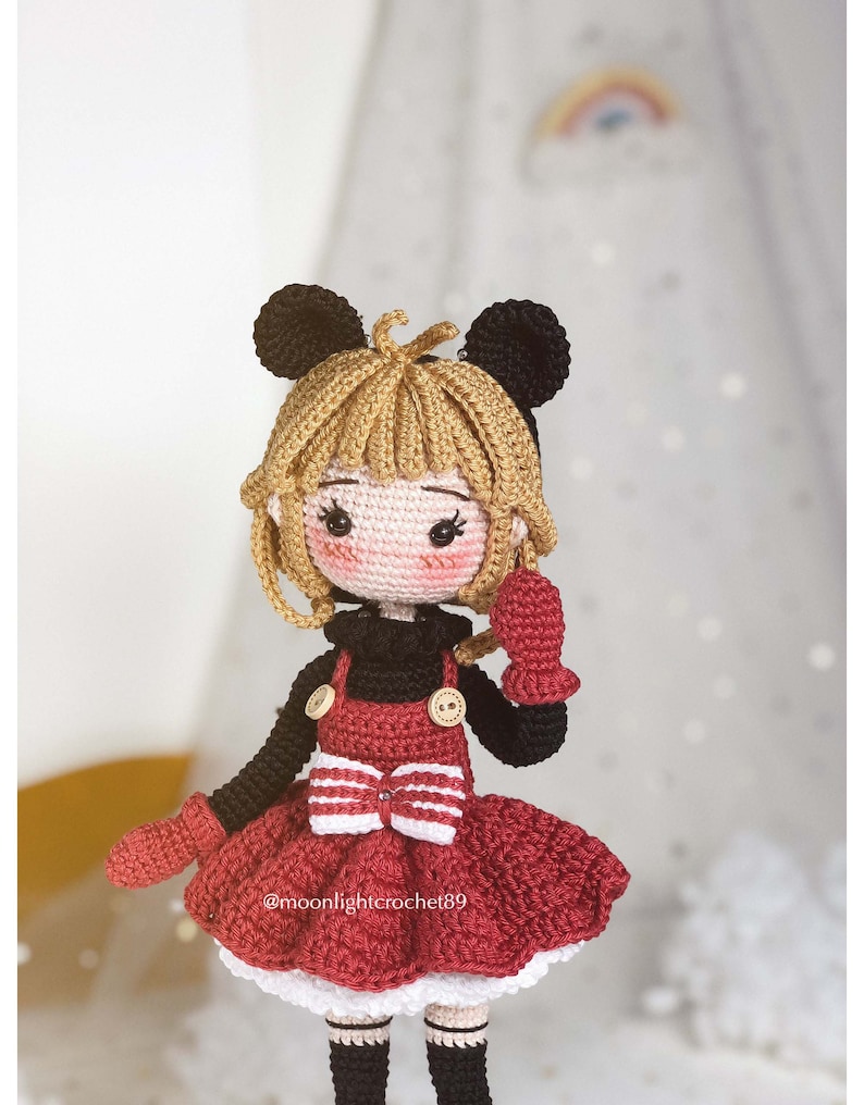 K&ouml;nnte beinhalten: Eine geh&auml;kelte Puppe mit blonden Haaren, die ein rotes Kleid mit einer wei&szlig;en Schleife, schwarze Schuhe und schwarze und rote F&auml;ustlinge tr&auml;gt. Die Puppe hat schwarze M&auml;useohren und einen schwarzen Pullover.