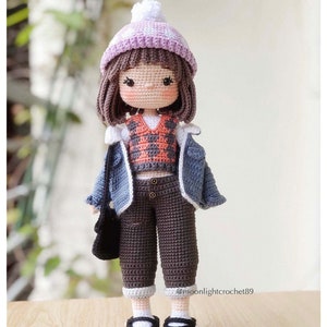 May include: A crocheted doll wearing a purple and white hat, a plaid waistcoat, a blue denim jacket, brown trousers, and black shoes. The doll is holding a black bag. @moonlightcrochet89