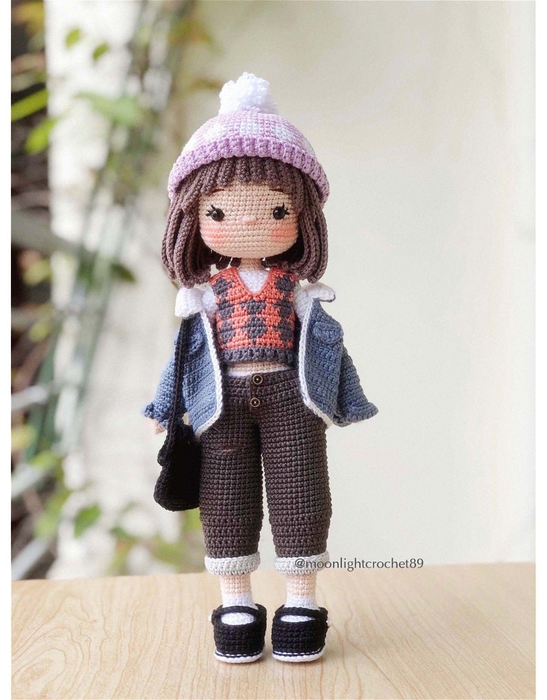 May include: A crocheted doll wearing a purple and white hat, a plaid waistcoat, a blue denim jacket, brown trousers, and black shoes. The doll is holding a black bag. @moonlightcrochet89