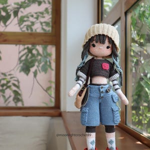 May include: A crocheted doll wearing a brown shirt, blue denim shorts, a beige hat, and red and brown shoes. The doll has blue yarn hair and is holding a brown bag. The doll is standing on a wooden surface. The text "@moonlightcrochet89" is visible in the image.
