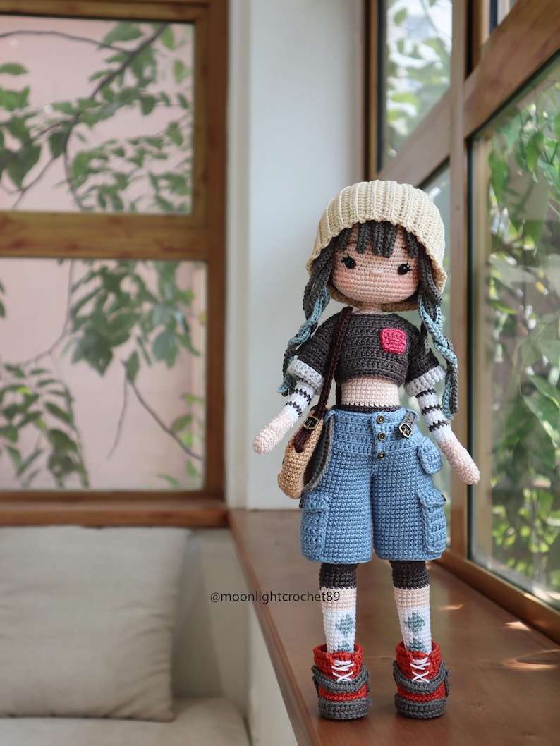 May include: A crocheted doll wearing a brown shirt, blue denim shorts, a beige hat, and red and brown shoes. The doll has blue yarn hair and is holding a brown bag. The doll is standing on a wooden surface. The text "@moonlightcrochet89" is visible in the image.
