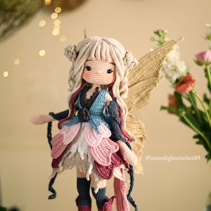 Peut inclure: Une poup&eacute;e de f&eacute;e au crochet faite &agrave; la main, avec des cheveux blonds et de grandes ailes beiges. La poup&eacute;e porte une robe &agrave; p&eacute;tales superpos&eacute;s roses, bleus et blancs. La poup&eacute;e a une expression douce et est entour&eacute;e de fleurs.
