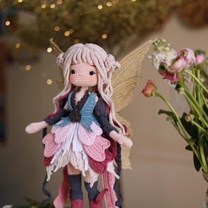 May include: A handmade crochet fairy doll with a light skin tone, long blonde hair, and delicate features. The doll wears a layered dress in shades of pink, white, and blue, with matching boots and wings. The doll is standing on a wooden surface.