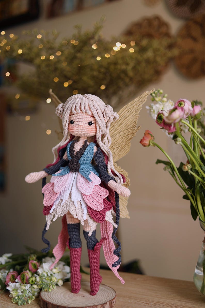 May include: A handmade crochet fairy doll with a light skin tone, long blonde hair, and delicate features. The doll wears a layered dress in shades of pink, white, and blue, with matching boots and wings. The doll is standing on a wooden surface.